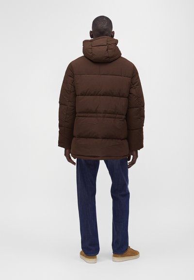 Homme debout de dos, portant une doudoune marron avec capuche, un jean bleu et des chaussures en daim beige sur un fond uni.
