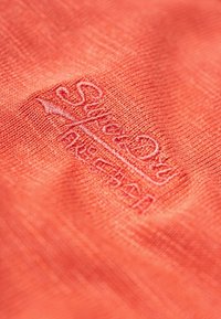 Tessuto color corallo con una trama sottile, caratterizzato da un logo ricamato in una tonalità contrastante. Design semplice e pulito.