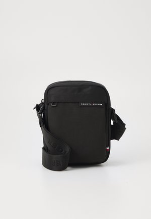 Sac bandoulière noir Tommy Hilfiger avec sangle réglable, poche zippée à l'avant et étiquette logo sur fond blanc uni.