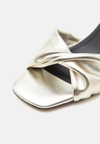 Κοντινό πλάνο μεταλλικού χρυσού σανδάλι με ανοιχτά δάχτυλα, στριμμένο λουράκι και τετράγωνο μπροστινό μέρος σε ανοιχτό φόντο.