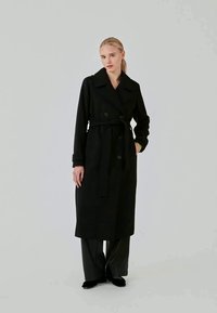 Abrigo negro de trench con un diseño de doble botonadura, cuello sobredimensionado y cinturón. Textura suave y mangas largas con puños.