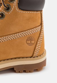 Tanfarbene Wildlederstiefel mit einem braunen Lederkragen, weißen Nähten und einem geprägten Baum-Logo. Die Sohle ist robust mit einer braunen Gummibeschichtung.