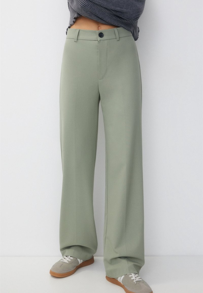 Personne portant un pantalon vert sauge taille haute et ample, avec un bouton noir, associé à des baskets beiges avec des lacets blancs.