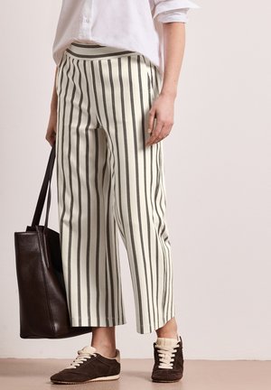 Parte inferiore del corpo di una persona che indossa pantaloni corti bianchi a righe, sneakers marrone scuro, camicia bianca e tiene una borsa tote marrone scuro.