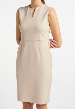 Beżowa sukienka bez rękawów do kolan z małym wycięciem na środku dekoltu, noszona przez osobę z rękami skrzyżowanymi za plecami.