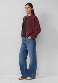 Burgunderfarbene Bomberjacke mit Reißverschluss vorne, kombiniert mit hellblauen weit geschnittenen Jeans. Beige Schuhe mit weißen Akzenten runden das Outfit ab.