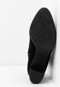Sort ruskindsko med indgraverede detaljer og logoer på sålen. Overdelen har en glat tekstur og en let forhøjet ryg for støtte.