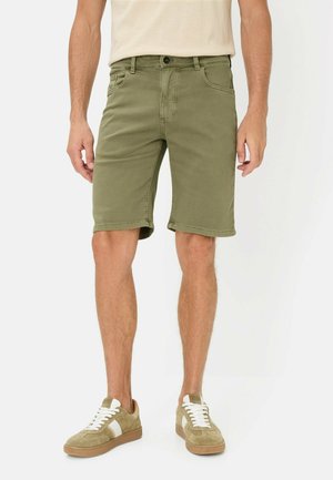 Mann trägt olivgrüne knielange Shorts und beige Turnschuhe mit weißen Streifen, steht vor einem weißen Hintergrund.