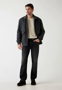 Veste grise avec un col rapproché, t-shirt clair, jean foncé et chaussures noires. La veste est dotée de poches avant et d'une fermeture éclair.