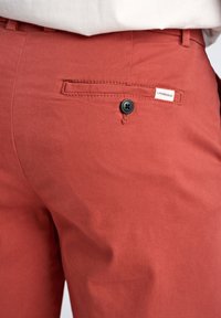 Pantalons en coton rouges avec une texture lisse, dotés d'une poche latérale, d'une fermeture à bouton noir et d'une petite étiquette blanche sur la taille.