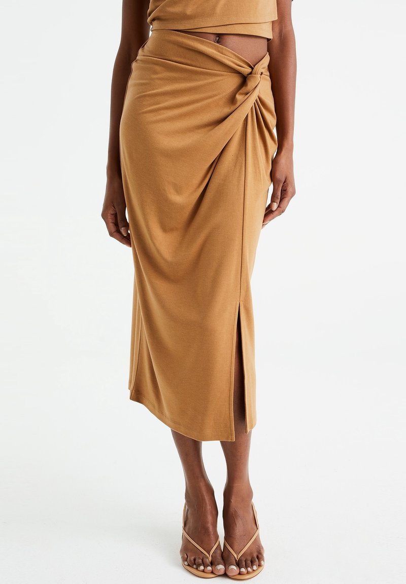 Hnědá sukně ke kolenům vyrobená z měkké tkaniny, s detailním uzlem na pase a bočním rozparkem pro větší pohyb.