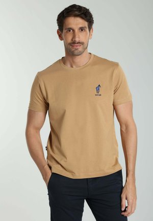 Homme portant un T-shirt beige à manches courtes avec un petit logo bleu de cheval sur la poitrine, debout avec une main dans la poche sur un fond uni.