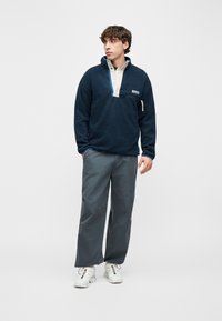 Νέος άνδρας που φοράει μπλέ φλις μπλούζα, γκρίζο παντελόνι με φαρδιά πόδια και λευκά αθλητικά παπούτσια, στέκεται μπροστά σε απλό φωτεινό φόντο κοιτάζοντας πλάγια.