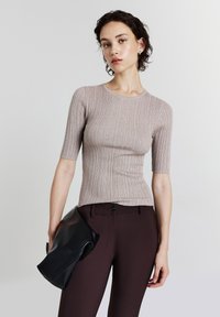 Top in maglia beige a coste con maniche corte, indossato con pantaloni sartoriali marrone scuro. Il modello tiene una pochette in pelle nera.