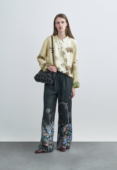 Blouse jaune à motifs floraux avec manches longues, associée à un pantalon noir à jambes larges orné d'accents floraux multicolores, portée avec un sac à main noir.