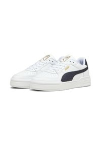 Sapatilhas brancas com um cabedal em pele texturizada, faixa lateral em azul marinho, marca em destaque e uma sola de borracha branca grossa. Detalhes em dourado na lingueta.
