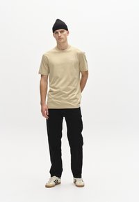 Beige bavlněné tričko s krátkými rukávy a malým bílým logem na hrudi, v kombinaci s černými kalhotami a bílými botami s černými pruhy.