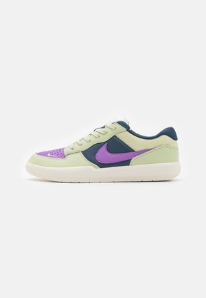 Baskets basses avec des panneaux crème, bleu foncé et violet, virgule Nike violette, semelle blanche et lacets crème sur fond blanc.
