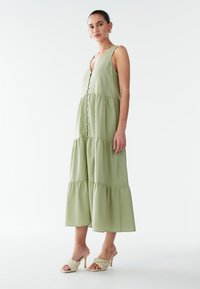 Lichtgroene maxi-jurk met laagjes, een V-hals, knopen aan de voorkant en mouwloos ontwerp. Gecombineerd met gevlochten mules met hak.