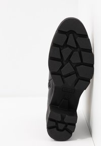 Semelle de botte noire avec motif de bande de roulement profond et robuste, taille 37, inclinée contre un mur et un sol blancs à angle droit.