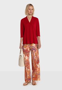 Haut rouge à col en V avec manches trois-quarts, associé à un pantalon large à motifs cachemire orange et un sac à main beige. Sandales pour compléter la tenue.