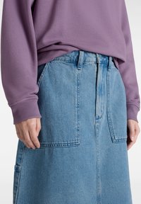 Sudadera morada con puños de canalé combinada con una falda de mezclilla azul que presenta grandes bolsillos frontales y un corte recto. Vista en primer plano del dobladillo.