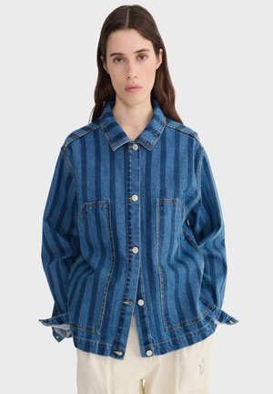 Žena s dlhými hnedými vlasmi, oblečená v modrej denimovej bunde s pásmi a prednými gombíkmi, a krémové nohavice s drobnou výšivkou.