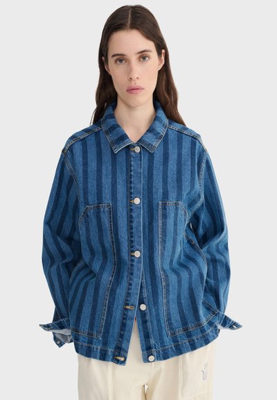 Femme aux longs cheveux bruns portant une veste en denim à rayures bleues avec des boutons sur le devant et un pantalon crème avec une petite broderie.