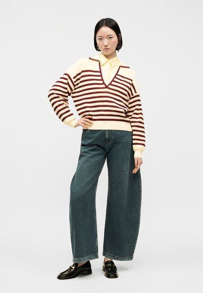 Femme debout, la main sur la hanche, portant un pull à col en V rayé crème et bordeaux, une chemise jaune à col, un jean à jambes larges et des mocassins noirs.