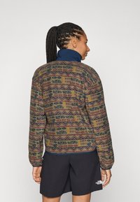 Fleecejakke med et mønsterdesign i jordfarver, med geometriske former i blå, bordeaux og gul, samt høj krave og elastiske manchetter.