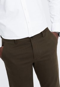 Pantaloni in cotone marrone a gamba dritta, con chiusura a bottone e tasche laterali, abbinati a una camicia bianca con bottoni.