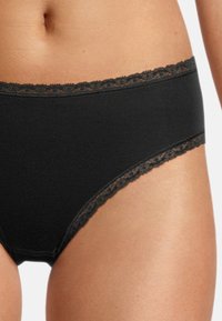 Slip in cotone nero con cintura piatta, caratterizzato da un delicato bordino in pizzo. Il tessuto ha una consistenza liscia e cuciture minime per garantire comfort.