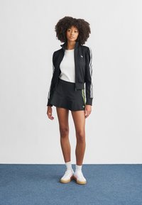 Veste de survêtement noire zippée avec des rayures blanches, associée à une jupe noire avec des accents verts. Le modèle porte des baskets blanches et des chaussettes basses.