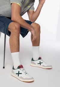 Białe i zielone trampki z teksturowanym wierzchem, z czerwonym logo, noszone z białymi skarpetkami w prążki i niebieskimi szortami dżinsowymi. Model siedzi na krześle.