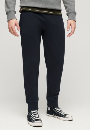 Pantaloni della tuta blu navy in morbido cotone, con polsini elastici alle caviglie e un piccolo logo ricamato sul davanti. Abbinati a sneakers nere.