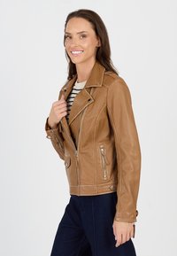 Braune Leder-Motorradjacke mit Reißverschlusstaschen, spitzem Kragen und Druckknopfdetails. Getragen über einem gestreiften Hemd und kombiniert mit dunklen Hosen.