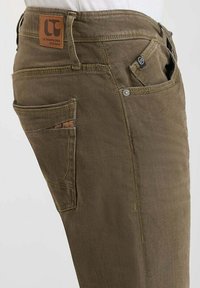 Pantalons en coton vert olive avec un patch en cuir, plusieurs poches et des coutures jaunes contrastantes. Ils disposent d'une taille ajustée et d'un tissu robuste.