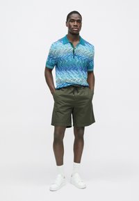 Camiseta tipo polo de manga corta con patrón ondulado azul, combinada con pantalones cortos verde oliva. Modelo lleva zapatillas blancas y calcetines. Fondo limpio.
