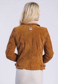 Eine Frau mit blonden Haaren steht mit dem Rücken zur Kamera, trägt eine braune Wildlederjacke mit einem Etikett und einen grauen Rock.