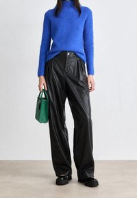 Person in blauer Rollkragenpullover und schwarzen weiten Lederhosen, hält eine grüne Handtasche und trägt schwarze klobige Schuhe.
