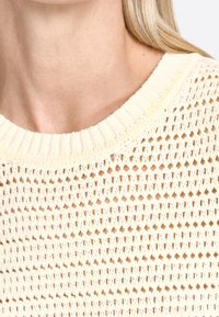 Sweter w kremowym kolorze, dziany, z szerokim ściągaczem przy dekolcie i otwartym wzorze, charakteryzujący się teksturowanym wyglądem i przewiewnym designem.