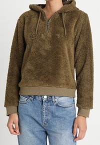 Fleecig huva-pullover i olivgrön, med en halvmaskerad dragkedja och ribbad nederkant. Mjuk textur och två justerbara dragsnören vid kragen.