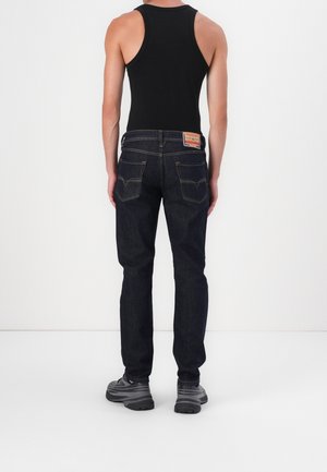Personne debout de dos portant un haut noir sans manches, un jean bleu foncé avec des poches arrière et des chaussures de sport grises sur fond blanc.