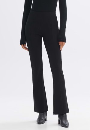 Personne portant un pantalon noir ajusté légèrement évasé et des bottines à talons noires, debout devant un fond clair uni.