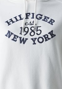 Felpa bianca con cappuccio con ricamo blu navy che recita "HILFIGER est. 1985 NEW YORK." Include lacci e una texture morbida e liscia.