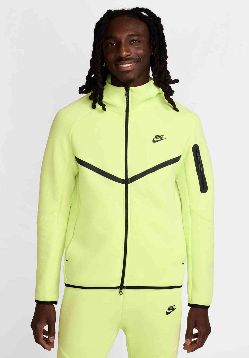 Chaqueta Nike amarilla neón con alto cuello, cierre frontal y acentos negros. El diseño presenta una franja diagonal y un bolsillo.