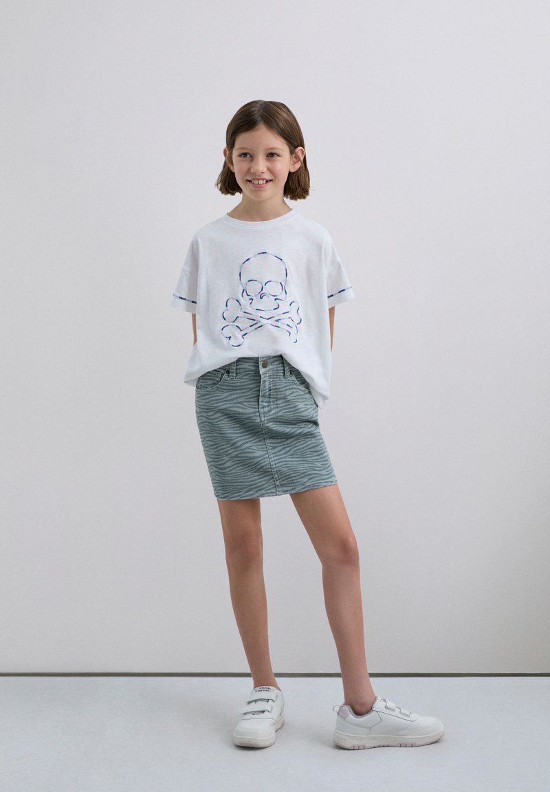 Fille souriante avec des cheveux bruns courts portant un t-shirt blanc surdimensionné avec un design de crâne, une jupe à motifs verts et des baskets blanches.