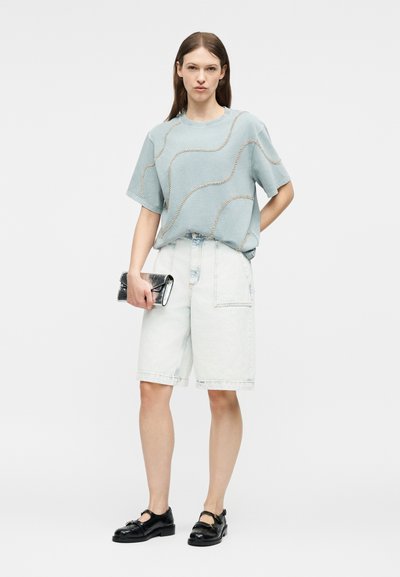Γυναίκα που φοράει κοντομάνικο μπλουζάκι γαλάζιου χρώματος με ραμμένα σχέδια, ανοιχτόχρωμο τζιν σορτς μέχρι τα γόνατα, μαύρα παπούτσια, κρατώντας ένα μαύρο πορτοφόλι.