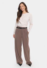 Svetlo bež kardigan z dolgimi rokavi in gumbi, v kombinaciji z visokim pasom in širokimi rjavimi hlačami ter črnimi čevlji s koničastim prstom.