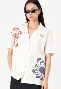 Chemise à manches courtes, crème, à boutons, en tissu léger avec des motifs floraux abstraits rouille et marine. Présente un accent logo sur la poitrine gauche.